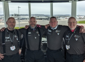 Four men in an airport control tower hugging and smiling